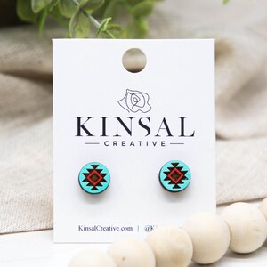 May include: A pair of round stud earrings with a turquoise background and a red and black geometric design. The earrings are displayed on a white card with the text "KINSAL CREATIVE" and the website address. The earrings are next to wooden beads.