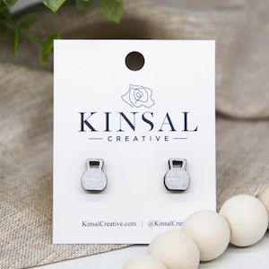 May include: A pair of silver-coloured wooden earrings shaped like kettlebells. The earrings are displayed on a white card with the text "Kinsal Creative" printed on it.