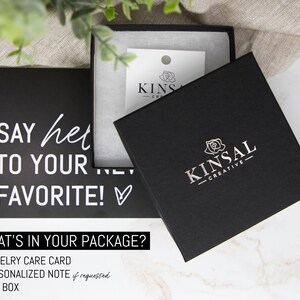 May include: A black gift box with a white rose and the text "KINSAL - CREATIVE" on the lid. The box is sitting on a white marble surface. The text "SAY hello TO YOUR FAVORITE!" is on a black card behind the box. The text "WHAT'S IN YOUR PACKAGE?" is below the card. A list of items included in the package is below the text: "JEWELRY CARE CARD", "PERSONALIZED NOTE if requested", "GIFT BOX".