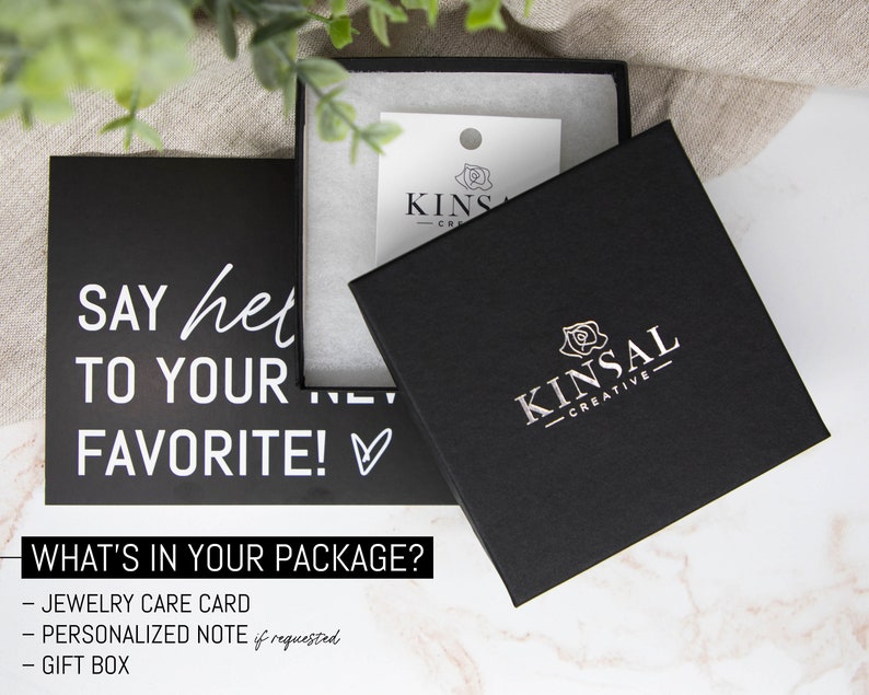 May include: A black gift box with a white rose and the text "KINSAL - CREATIVE" on the lid. The box is sitting on a white marble surface. The text "SAY hello TO YOUR FAVORITE!" is on a black card behind the box. The text "WHAT'S IN YOUR PACKAGE?" is below the card. A list of items included in the package is below the text: "JEWELRY CARE CARD", "PERSONALIZED NOTE if requested", "GIFT BOX".