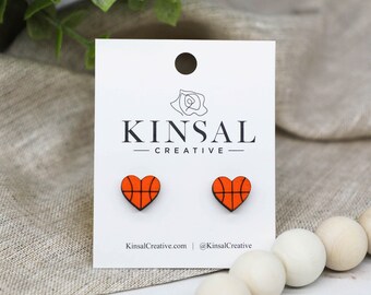 May include: A pair of heart-shaped earrings with a basketball design. The earrings are made of wood and are painted orange and black. The earrings are on a white card with the text "Kinsal Creative" printed on it.