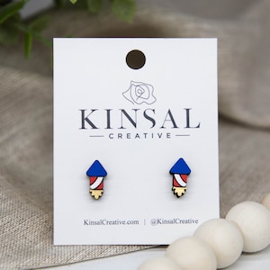 May include: A pair of small, wooden stud earrings shaped like red, white, and blue fireworks. The earrings are on a white card with the text "Kinsal Creative" printed on it.