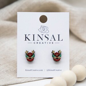 May include: A pair of wooden earrings featuring a red and green dragon design. The earrings are on a white card with the text "Kinsal Creative" printed on it.