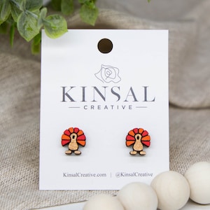 May include: A pair of wooden turkey earrings with a red, orange, and yellow design. The earrings are on a white card with the text "Kinsal Creative" printed on it.