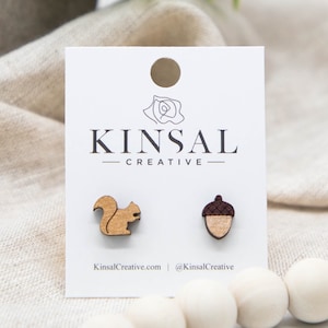 May include: A pair of wooden earrings featuring a squirrel and an acorn. The earrings are brown and have a natural wood finish. The earrings are displayed on a white card with the text "Kinsal Creative" printed on it.