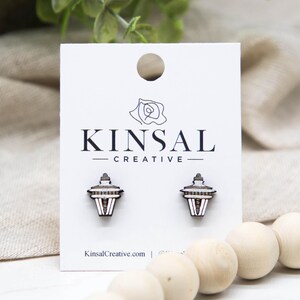 May include: Wooden stud earrings featuring a miniature lantern design. The earrings are displayed on a white card with the text "KINSAL CREATIVE" and the website address. The earrings have a brown, white, and black color scheme.