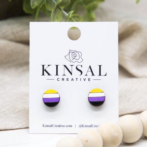 May include: A pair of round stud earrings featuring the non-binary pride flag design. The earrings have a black base with horizontal stripes of yellow, white, purple, and black. The earrings are displayed on a white card with the text "KINSAL CREATIVE".