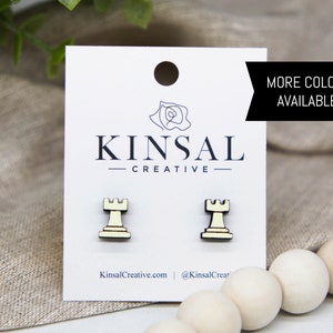 May include: A pair of wooden earrings shaped like chess rooks. The earrings are a light yellow color with black accents. The earrings are displayed on a white card with the text "Kinsal Creative" and "More Colors Available!"