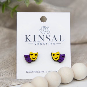 May include: A pair of wooden earrings featuring a purple and yellow painted design of two theater masks. The earrings are displayed on a white card with the text "Kinsal Creative" printed on it.