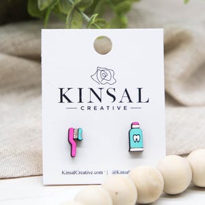 May include: Wooden earrings with a toothbrush and toothpaste design. The toothbrush is pink with a green brush, and the toothpaste is blue with a pink cap. The earrings are on a white card with the "Kinsal Creative" logo.