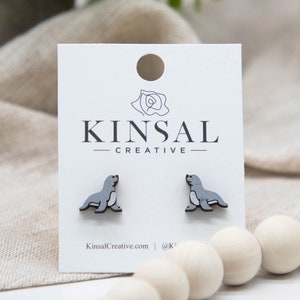 Puede incluir: Pendientes de botón de madera con diseño de foca gris y negra, presentados en una tarjeta blanca con el texto "KINSAL CREATIVE". La tarjeta tiene un agujero en la parte superior. Fondo neutro con cuentas de madera.