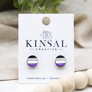 May include: A pair of round stud earrings featuring the asexual pride flag design. The earrings are black, gray, white, and purple. The earrings are displayed on a white card with the text "KINSAL CREATIVE".