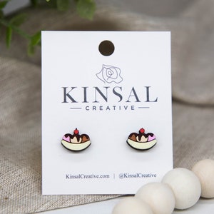 Puede incluir: Un par de pendientes de madera con forma de banana split con helado de chocolate, fresa y vainilla, coronado con una cereza. Los pendientes están en una tarjeta blanca con el texto "Kinsal Creative".