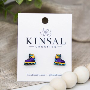 Op de afbeelding: Een paar rolstoel-oorbellen in paars, geel en blauw. De oorbellen zijn gemaakt van hout en ongeveer 2,5 cm groot. De oorbellen liggen op een witte kaart met de tekst "Kinsal Creative" erop gedrukt.