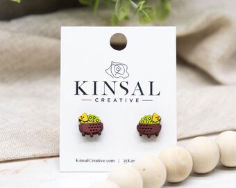 Può includere: Orecchini a bottone in legno con design di succulente. Gli orecchini raffigurano un piccolo vaso con una succulenta verde e gialla. Gli orecchini sono su una carta bianca con la scritta "KINSAL CREATIVE".