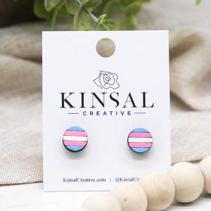 May include: Wooden stud earrings featuring the transgender pride flag design. Each earring has a blue top and bottom, a white center stripe, and a pink stripe. The earrings are displayed on a white card with the "KINSAL CREATIVE" logo and website address.