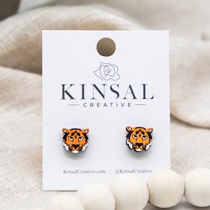Pode incluir: Um par de brincos de pressão com a forma de um rosto de tigre em um cartão branco com o texto "Kinsal Creative".