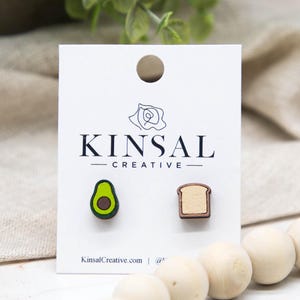 May include: Wooden stud earrings featuring an avocado and a slice of toast. The avocado earring is green with a brown pit, and the toast earring is light brown. The earrings are displayed on a white card with the text "KINSAL CREATIVE".