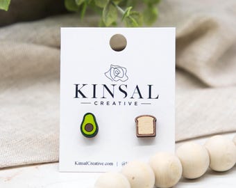 May include: Wooden stud earrings featuring an avocado and a slice of toast. The avocado earring is green with a brown pit, and the toast earring is light brown. The earrings are displayed on a white card with the text "KINSAL CREATIVE".