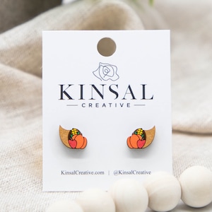 May include: A pair of wooden earrings featuring a cornucopia design with pumpkins, corn, and leaves. The earrings are painted in shades of orange, yellow, red, and green. The earrings are displayed on a white card with the text "Kinsal Creative" printed on it.