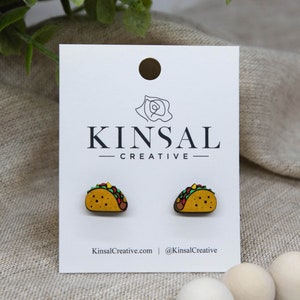 May include: A pair of wooden taco earrings. The earrings are yellow with green, red, and brown details. The earrings are on a white card with the text "Kinsal Creative" printed on it.