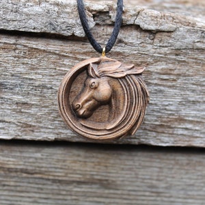 May include: A wooden horse head pendant necklace with a brown finish. The pendant is round and features a detailed carving of a horse's head. The necklace is made of black cord.