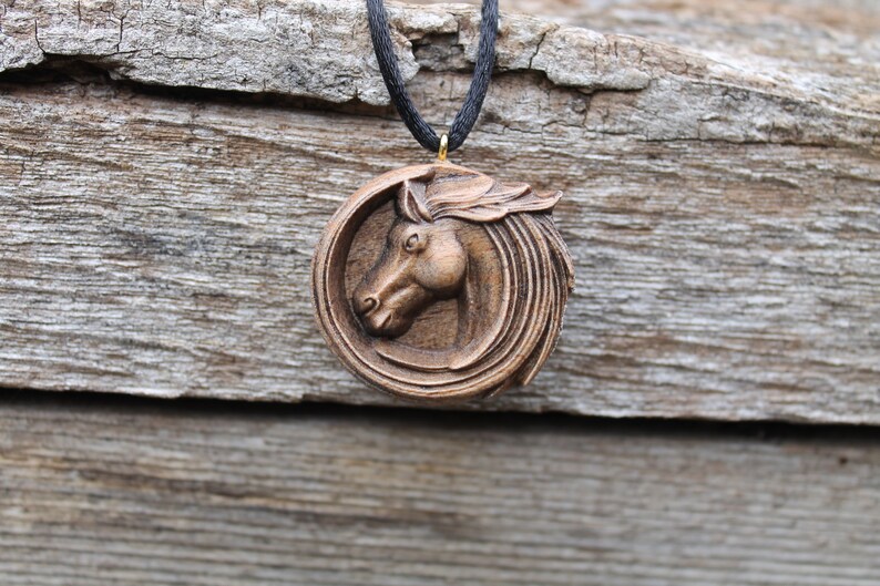 May include: A wooden horse head pendant necklace with a brown finish. The pendant is round and features a detailed carving of a horse's head. The necklace is made of black cord.