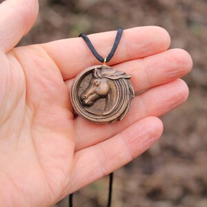 May include: A wooden horse head pendant necklace with a brown cord. The pendant is round and features a detailed carving of a horse's head.