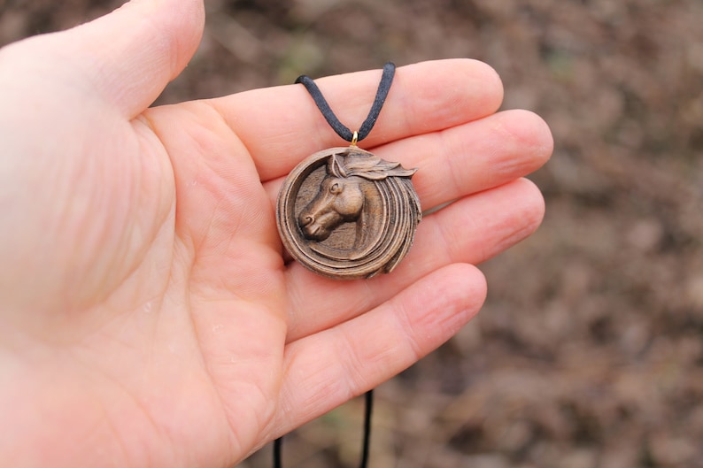 May include: A wooden horse head pendant necklace with a brown cord. The pendant is round and features a detailed carving of a horse's head.