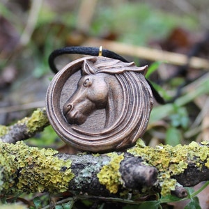 May include: A wooden pendant featuring a carved horse head with a black cord. The pendant is on a branch with green moss.