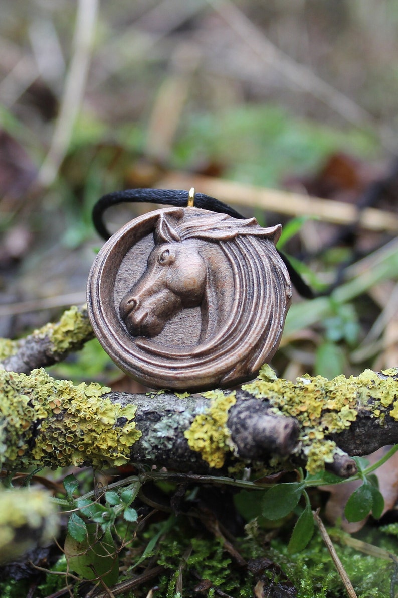 May include: A wooden pendant featuring a carved horse head with a black cord. The pendant is on a branch with green moss.