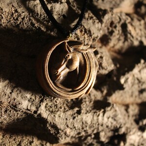 May include: A wooden pendant with a carved horse head design. The pendant is on a black cord and is hanging from a rough, brown surface.