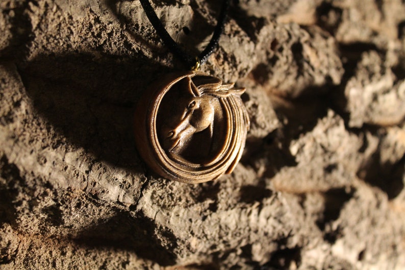 May include: A wooden pendant with a carved horse head design. The pendant is on a black cord and is hanging from a rough, brown surface.
