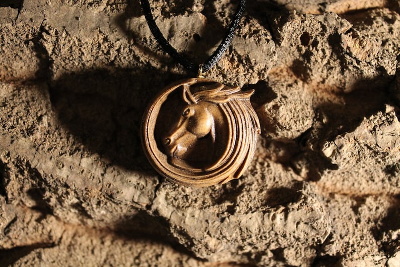 May include: A wooden pendant necklace featuring a carved horse head design. The pendant is round and has a dark brown color. The horse's mane is flowing and detailed.