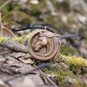 May include: A wooden horse head pendant necklace with a brown cord. The pendant is carved with intricate details and has a gold-colored finish.