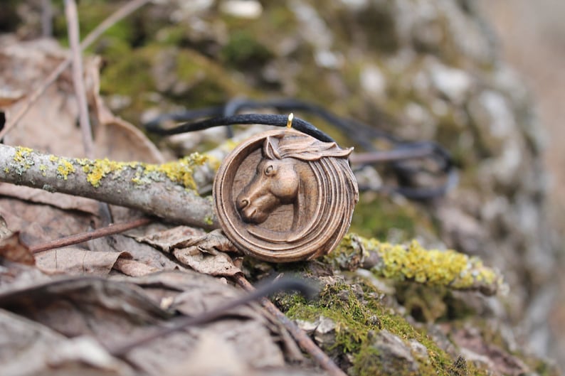 May include: A wooden horse head pendant necklace with a brown cord. The pendant is carved with intricate details and has a gold-colored finish.
