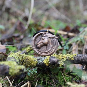 May include: A wooden pendant with a carved horse head design. The pendant is brown and has a black cord attached to it. The pendant is lying on a branch covered in green moss.