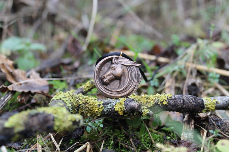 May include: A wooden pendant with a carved horse head design. The pendant is brown and has a black cord attached to it. The pendant is lying on a branch covered in green moss.