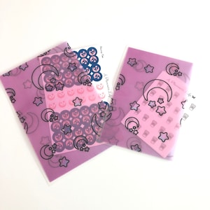 May include: Two clear plastic pouches with a purple background and a repeating pattern of black crescent moons and stars. The pouches contain sheets of stickers with a pink background and a repeating pattern of crescent moons and stars. The stickers feature a coffee cup design.
