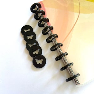 May include: Black planner discs with butterfly cutouts and a silver binding. The discs are arranged in a row, with some loose discs scattered nearby. The background is a light-colored planner page with watercolor-like designs.