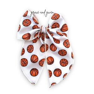 May include: A white hair bow with a basketball pattern. The bow is tied in a large, symmetrical knot with two long tails.