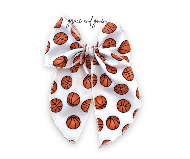May include: A white hair bow with a basketball pattern. The bow is tied in a large, symmetrical knot with two long tails.