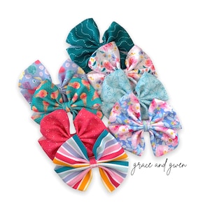 May include: A set of eight colorful hair bows with various patterns, including floral, geometric, and abstract designs. The bows are made of fabric and are tied with a ribbon. The text "grace and gwen" is visible in the bottom right corner of the image.