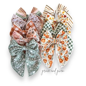May include: A set of 8 fall-themed hair bows in various patterns, including gingham, floral, and pumpkin prints. The bows are made with a variety of fabrics in shades of green, orange, brown, and white. The bows are arranged in a stack, with the text "grace and given" visible at the bottom.
