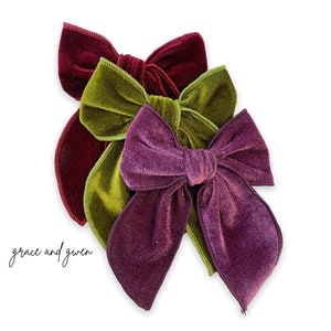 May include: Three velvet hair bows in shades of burgundy, green, and purple. The bows are arranged in a stack, with the burgundy bow on the bottom, the green bow in the middle, and the purple bow on top.