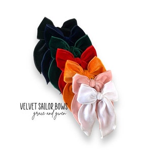 May include: A set of six velvet sailor bows in various colors, including navy blue, green, red, orange, pink, and white. The bows are arranged in a stack, with the largest bow on the bottom and the smallest bow on top. The text "VELVET SAILOR BOWS grace and gwen" is printed below the bows.