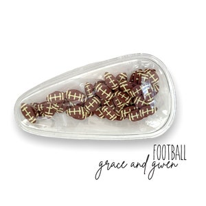 May include: A clear plastic football-shaped container filled with small brown and white football-shaped charms. The text "FOOTBALL grace and given" is written below the container.