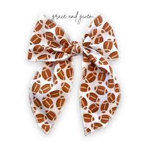 May include: A white hair bow with a brown football pattern. The bow is tied in a large, symmetrical knot.