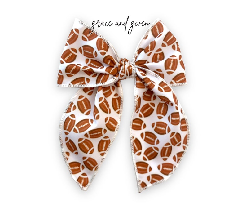 May include: A white hair bow with a brown football pattern. The bow is tied in a large, symmetrical knot.