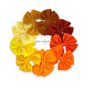 May include: A circular arrangement of ten fabric hair bows in a gradient of warm colors, ranging from pale yellow to deep brown and red-orange. Each bow features a textured, ribbed fabric and a central knot. The text grace and gwen is visible.